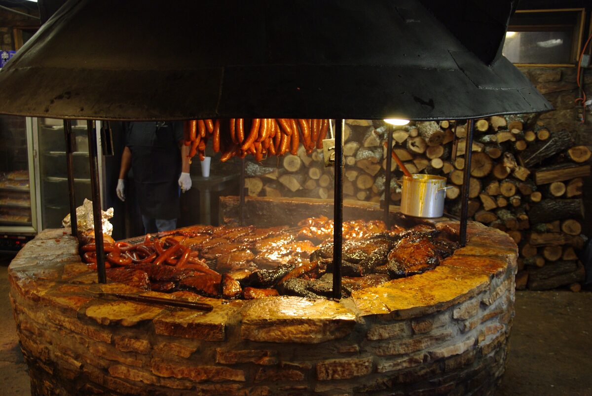 A round stone barbecue pit with ribs and sausages over the fire and split firewood stacked behind it