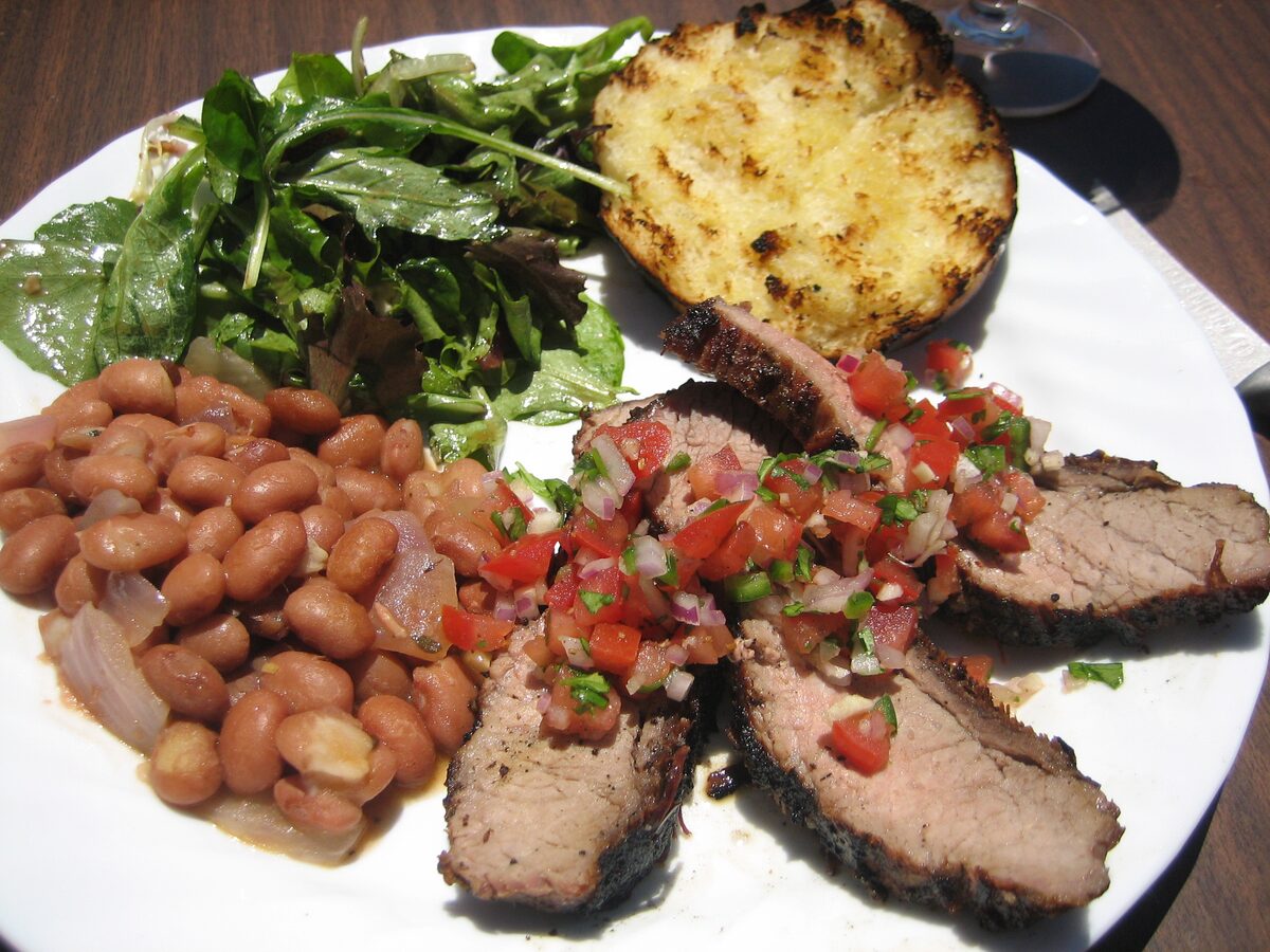 Sliced Santa Maria-style tri-tip with salsa, pink beans, salad and garlic bread on a white plate