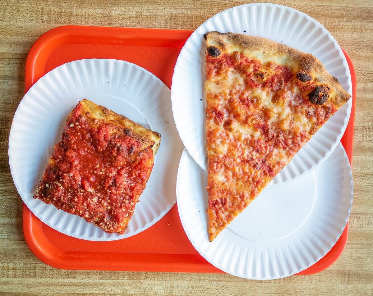 A plain New York slice and a square slice on paper plates on an orange tray at a slice shop