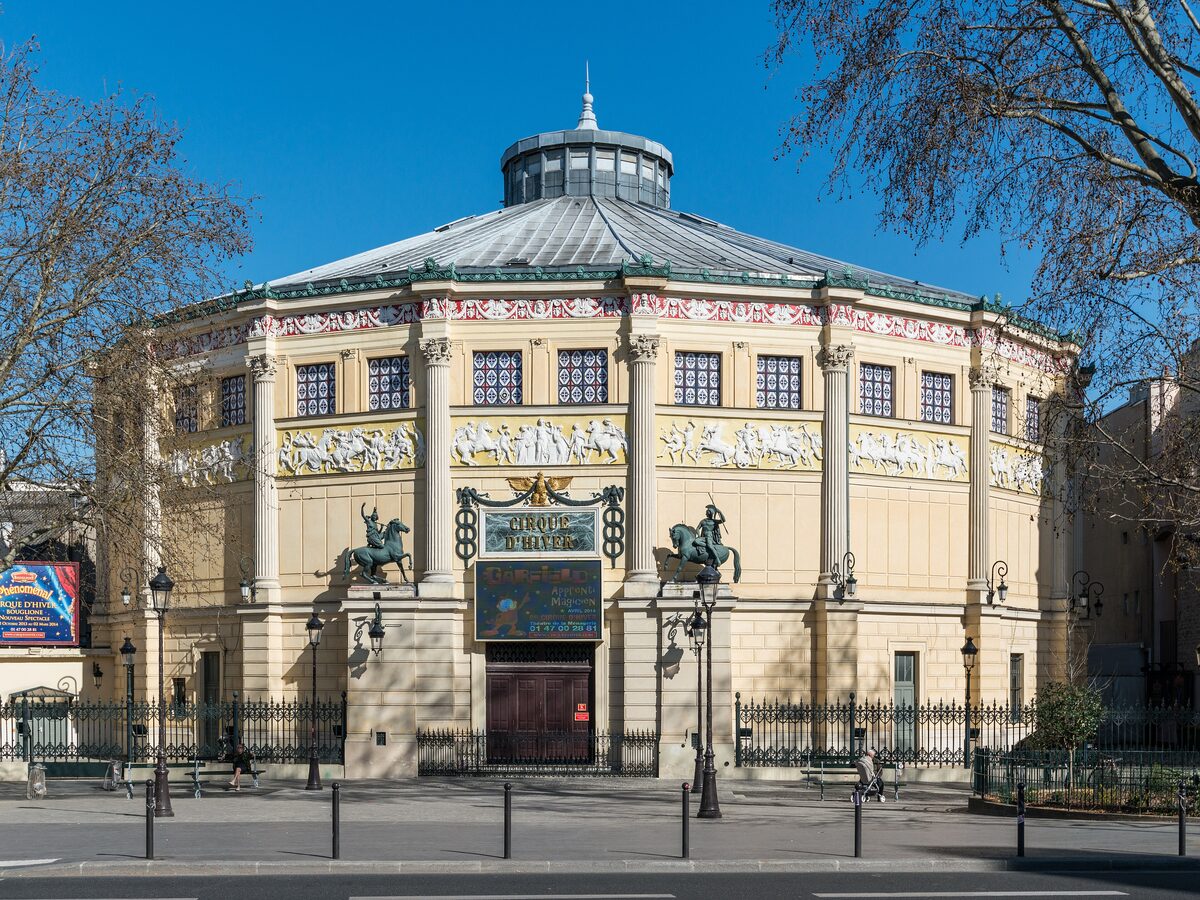 The round stone Cirque d'Hiver building in Paris, with bronze horse statues at the entrance and a glass-topped roof