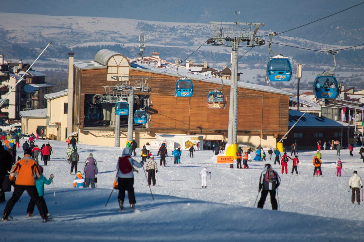 Skiers at the Bansko gondola base station, with gondola cabins and a lift tower overhead