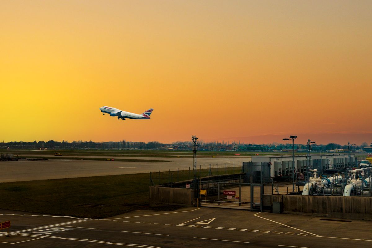 A passenger jet climbing away from the runway at Heathrow against an orange sunset sky