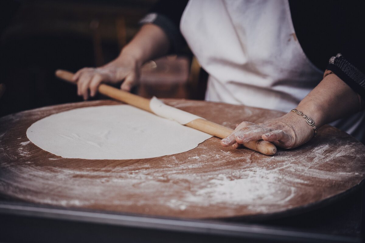 A woman rolling out a thin round of yufka dough with a long wooden rolling pin on a floured board