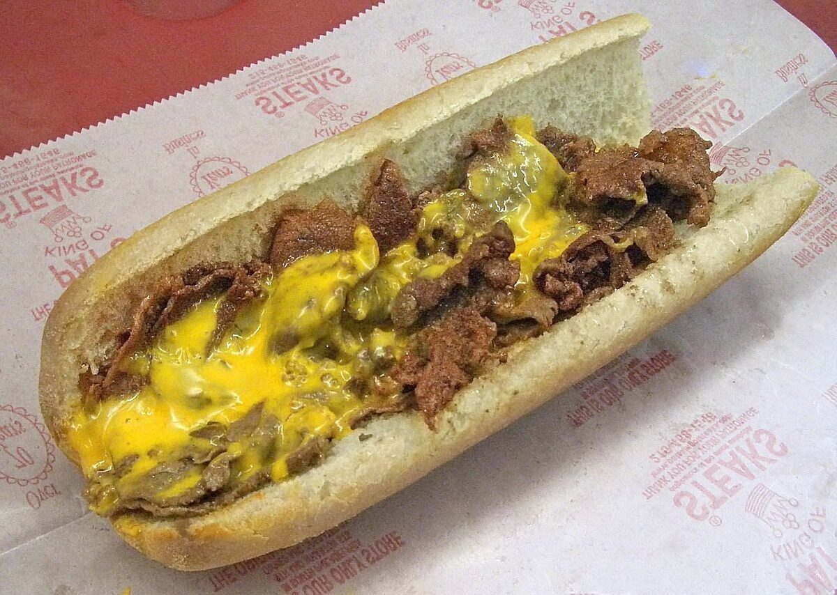 A cheesesteak on a long roll, filled with chopped beef and melted yellow cheese, on Pat's printed paper