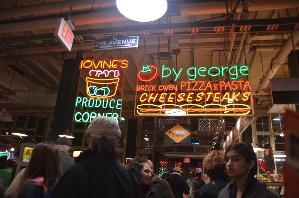 Neon signs for produce, brick oven pizza and cheesesteaks above the crowded stalls inside Reading Terminal Market