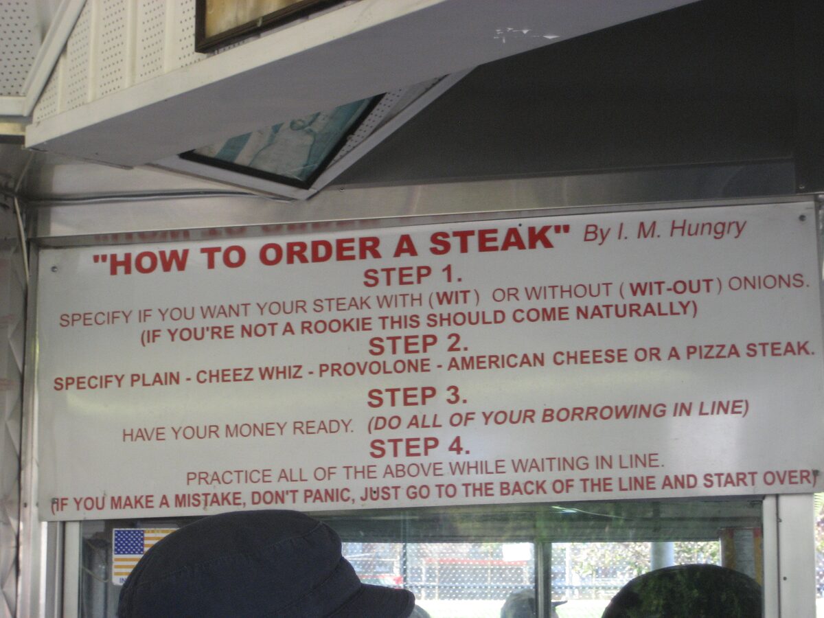 A sign at a cheesesteak window titled How to Order a Steak: say wit or wit-out onions, pick your cheese, have your money ready