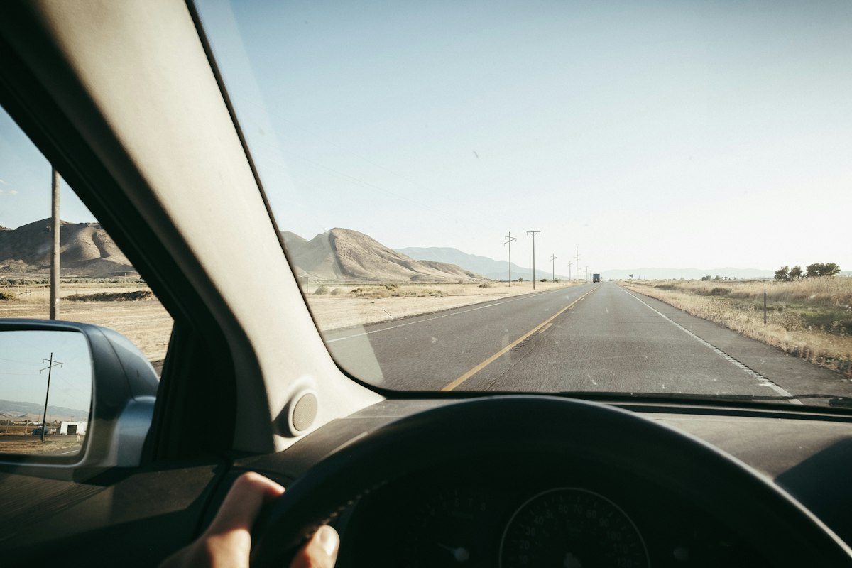 Driver's seat view with a hand on the wheel and an open desert highway ahead in Utah