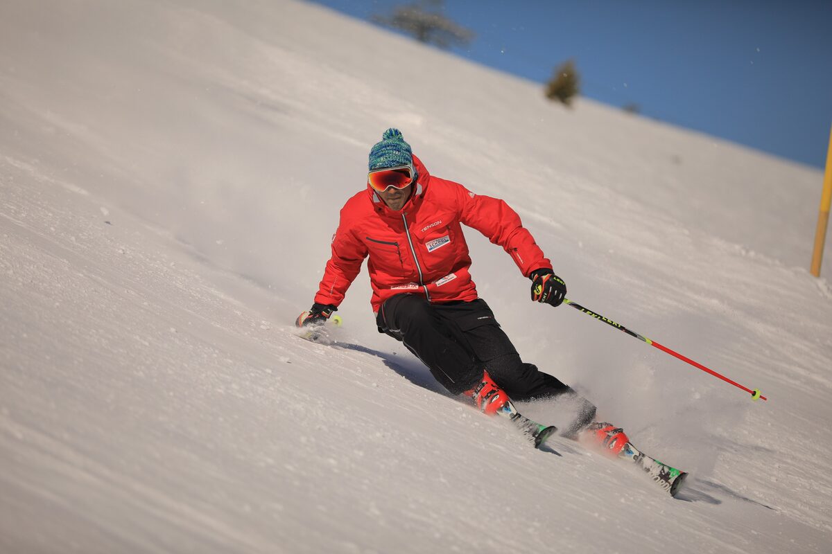 A ski instructor in a red jacket carving a turn on a sunny, groomed slope at Bansko