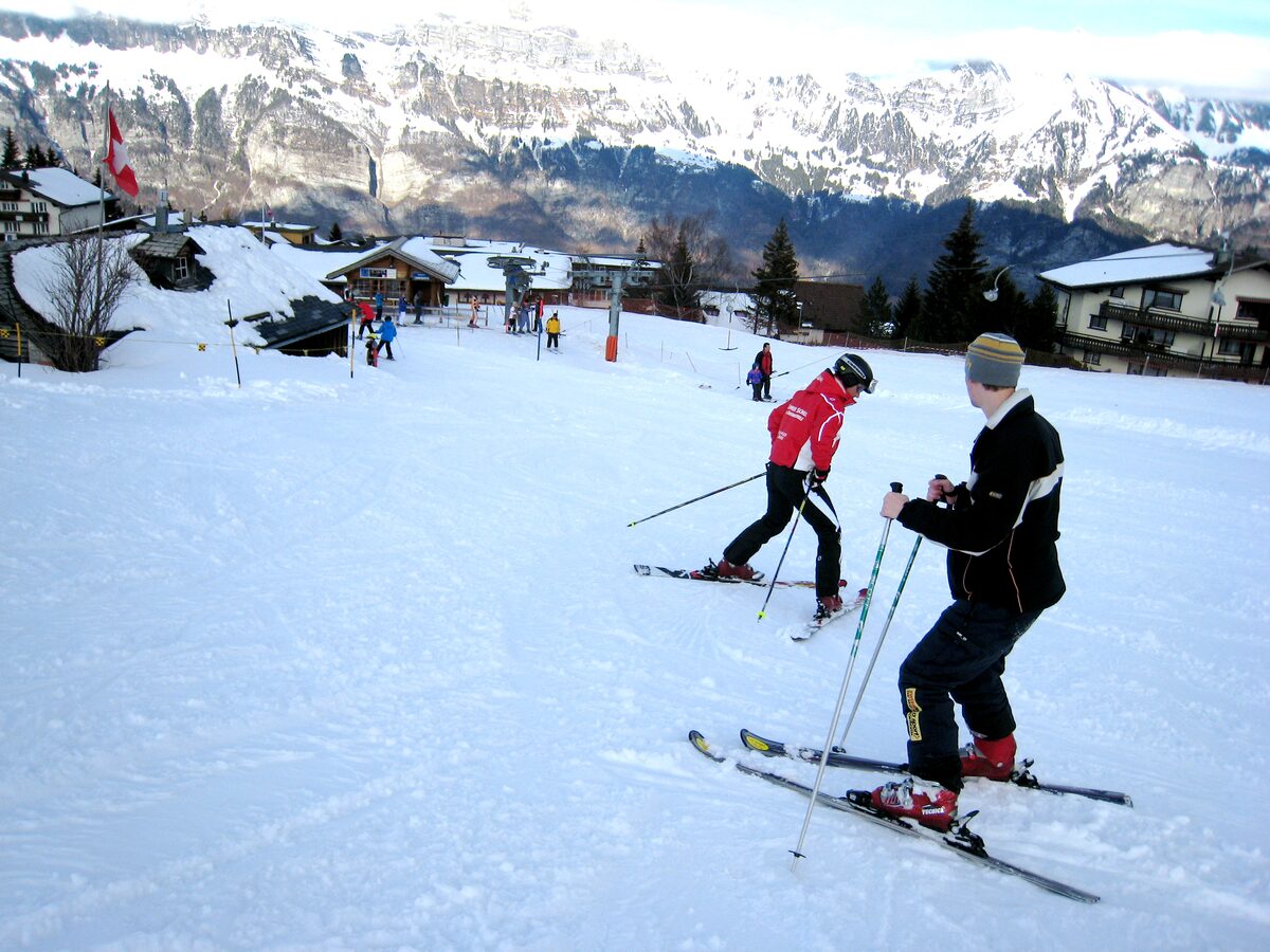 Two skiers stopped on a wide, gentle snowy slope above a village, one in a red jacket and one in black