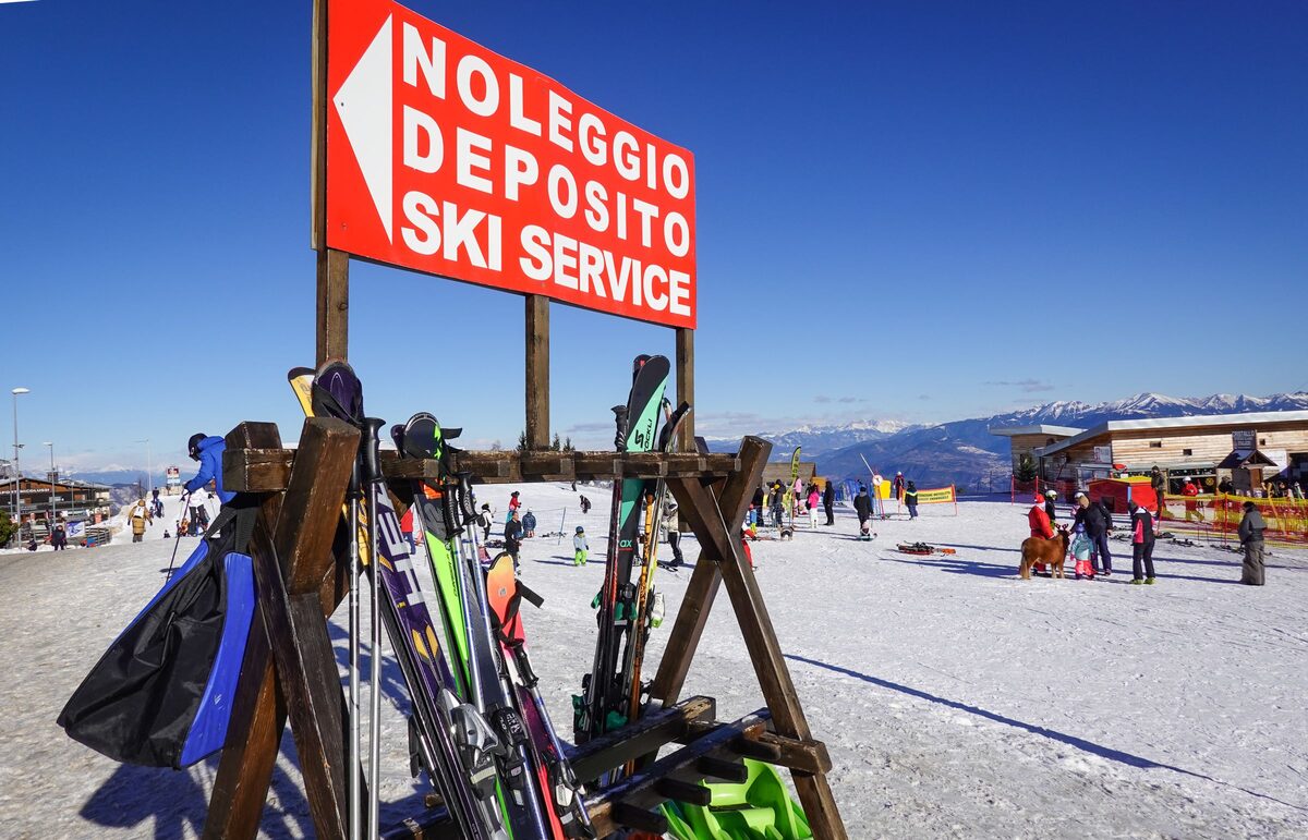 Skis and poles in a wooden rack beneath a Noleggio Deposito Ski Service sign, the Italian for rental and storage, on a sunny snowy slope