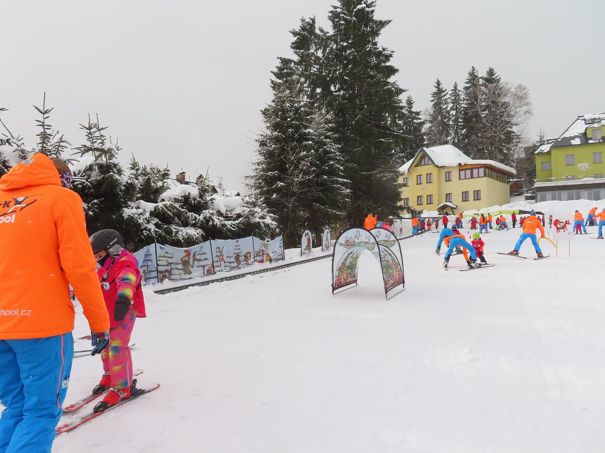 A ski instructor in an orange jacket guiding a small child on skis, with other ski school groups behind on the nursery slope
