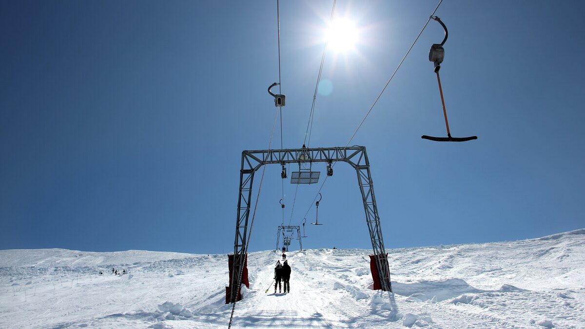 A T-bar drag lift on a snowy slope under a bright sun, with two skiers at the bottom