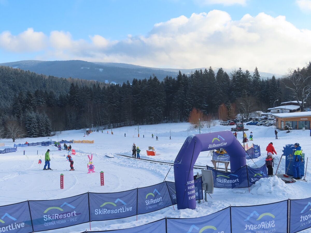 A snowy beginner area with a magic carpet, children and instructors on a gentle slope at Janske Lazne