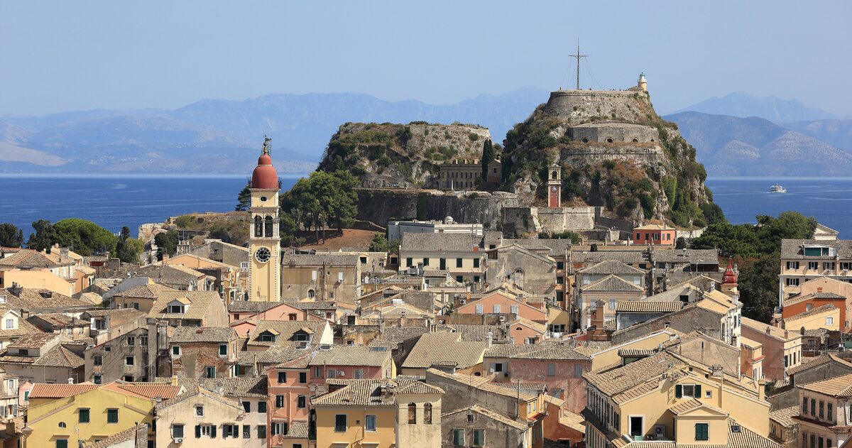 The Old Fortress rising above the terracotta roofs and bell tower of Corfu Old Town, with the Albanian coast behind