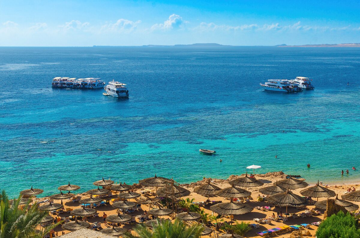 Turquoise water and coral reef at Ras Um Sid near Sharm El Sheikh, with dive boats and a beach of straw umbrellas