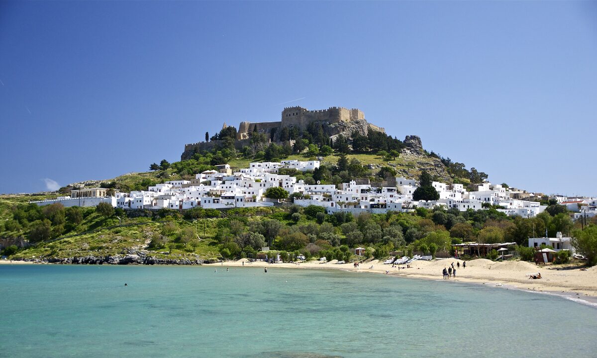The white houses of Lindos below its hilltop acropolis, above a sandy bay with calm turquoise water