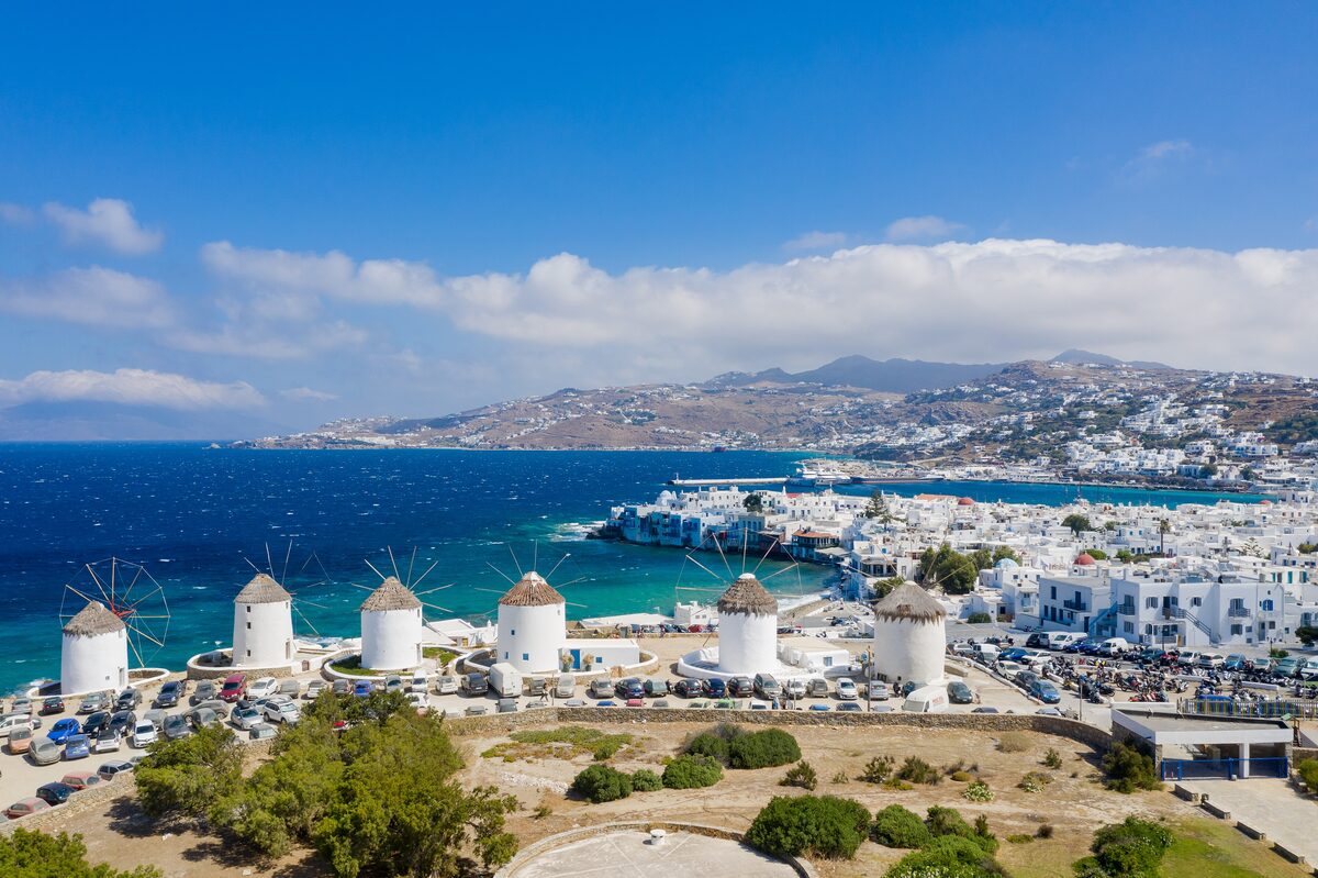 A row of white windmills above the sea in Mykonos Town, with Little Venice and the harbor behind