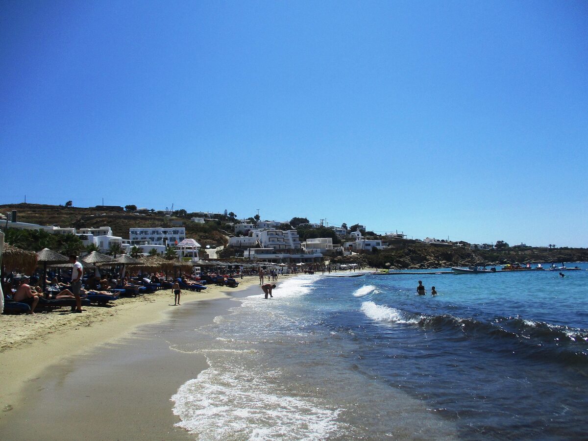A sandy beach with sunbeds and breaking waves at Platys Gialos on Mykonos, white houses on the hill behind