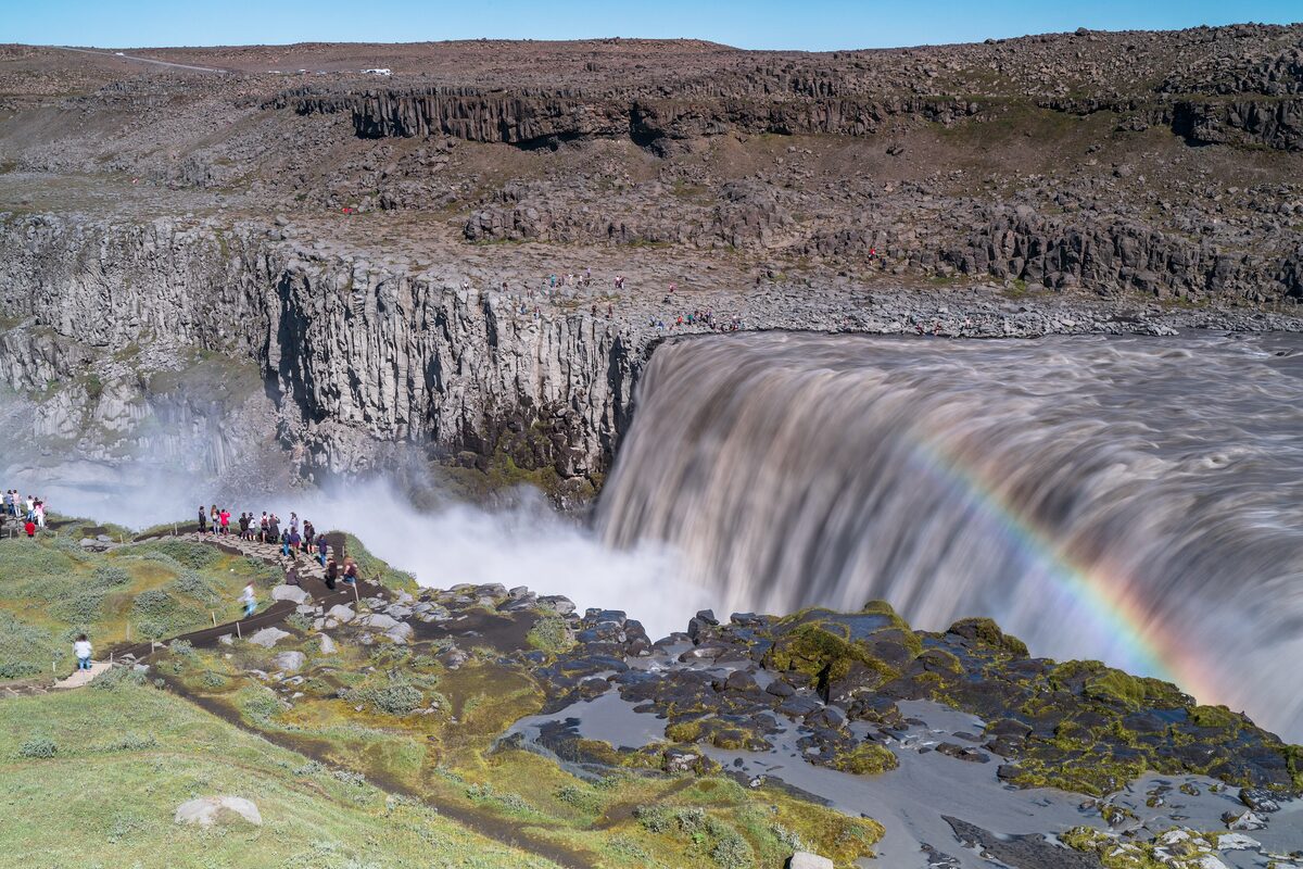 Dettifoss in daylight, the broad brown glacial river pouring over a basalt edge with a rainbow in the spray and visitors on the rim path
