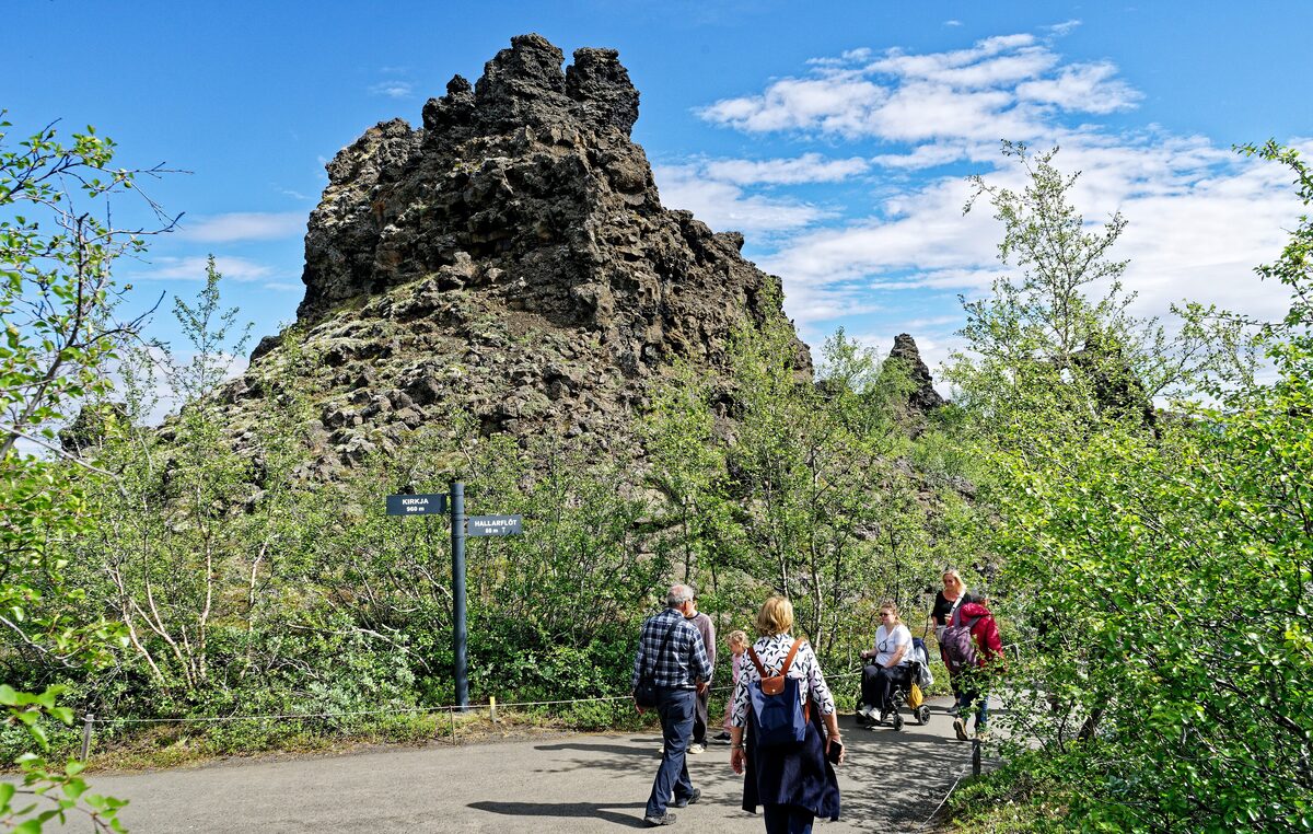 A dark lava pillar at Dimmuborgir with walkers on the path below it, green birch trees around