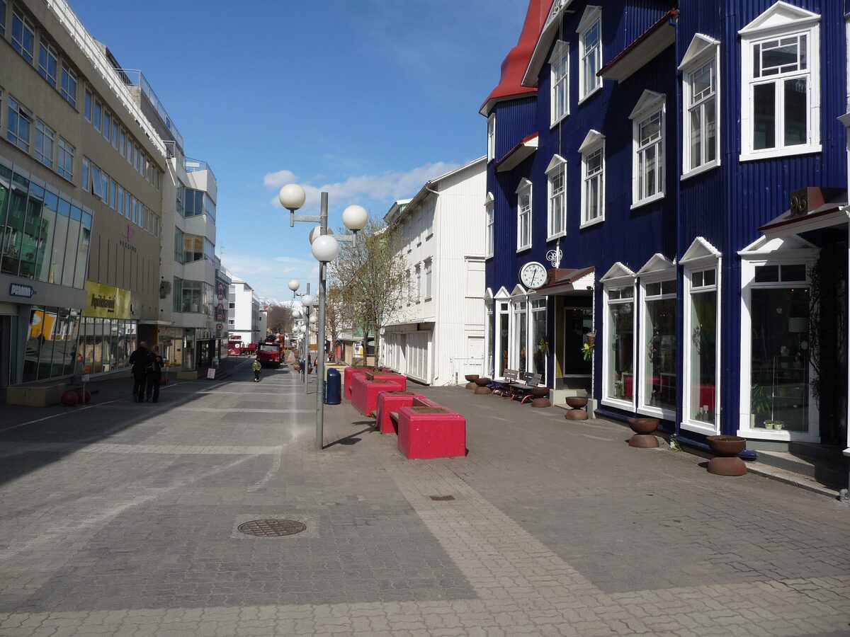 The pedestrian stretch of Hafnarstræti in central Akureyri on a sunny day, with a dark blue timber building with a red roof on the right and globe street lamps down the middle