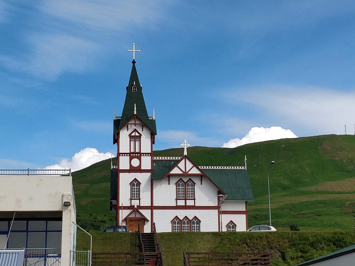 The white wooden Húsavík church with a green-roofed spire and red trim, on a grassy rise with a green hill and blue sky behind