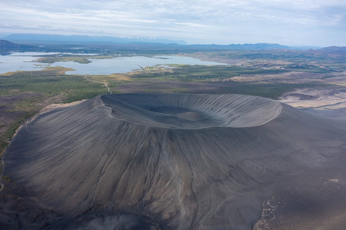Aerial view of the Hverfjall tephra ring, a round dark crater with ash slopes, with Lake Mývatn and green lowlands behind