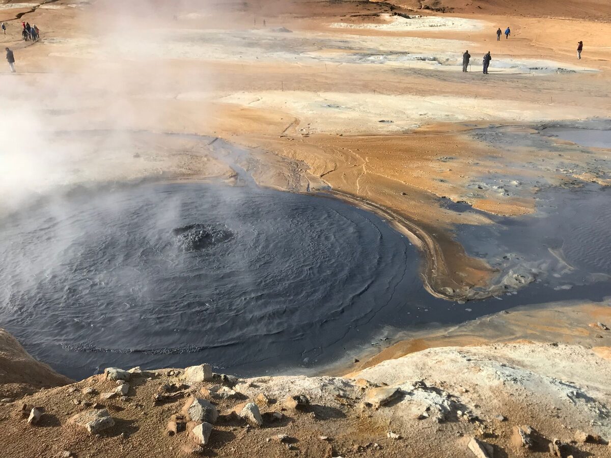A steaming grey mud pot in the Hverir geothermal field, with orange and yellow sulfur-stained ground and a few visitors on the paths beyond