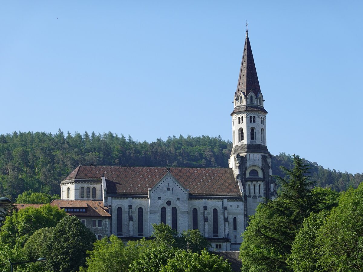 The Basilica of the Visitation above Annecy, a pale stone church with a tall pointed spire and a patterned brown-tiled roof, in front of a wooded hill