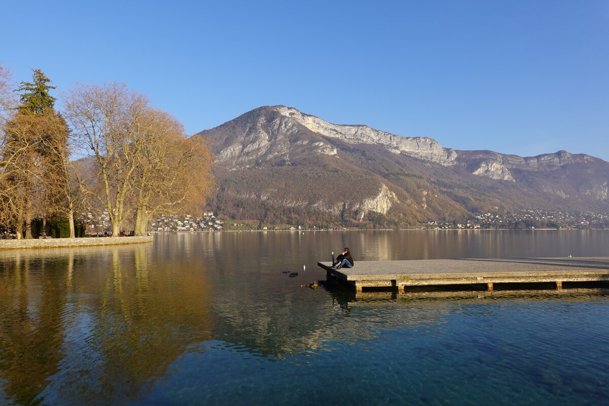 A person sitting at the end of a flat jetty on the calm Annecy lake, with autumn plane trees on the shore and a limestone mountain across the water