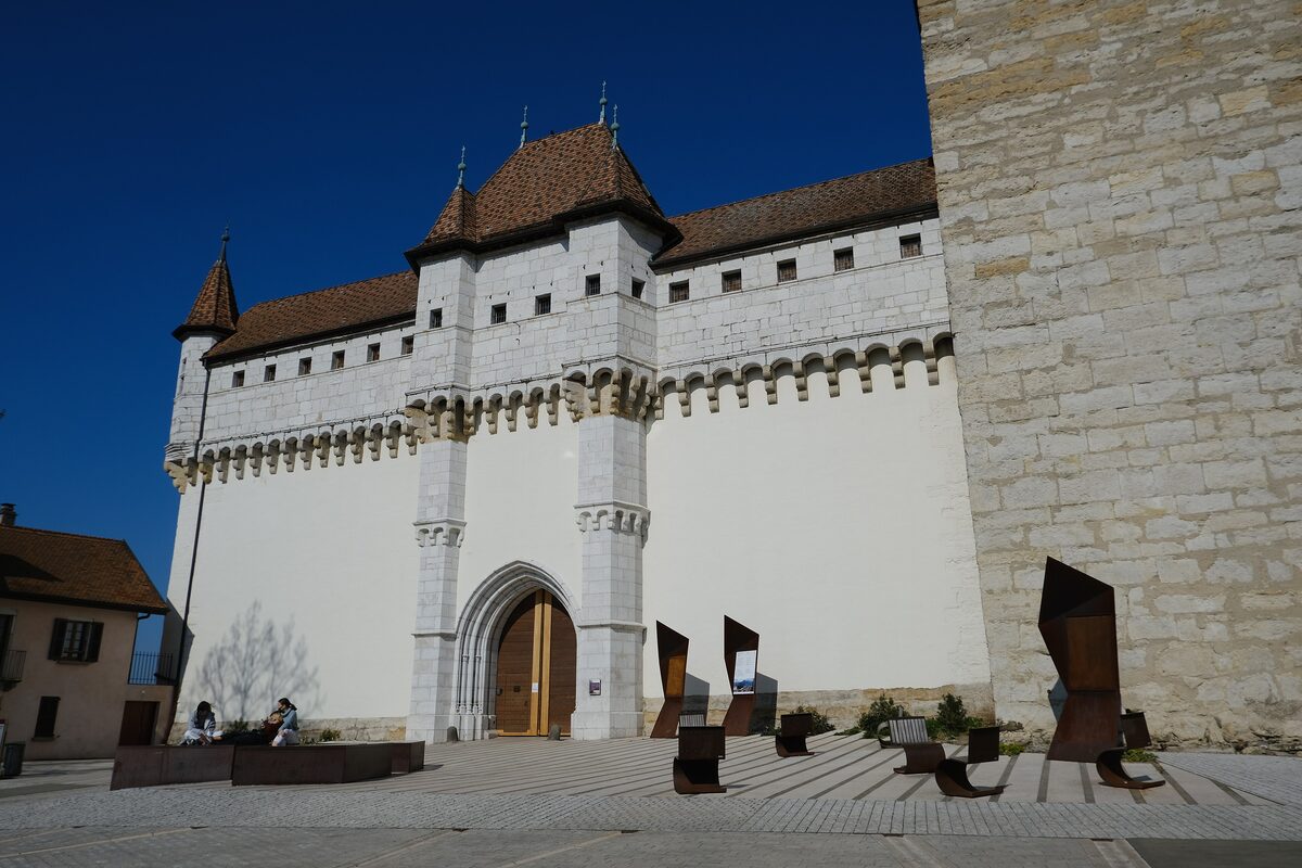 The white facade of the Château d'Annecy with pointed towers and an arched doorway, and dark metal sculptures on the forecourt