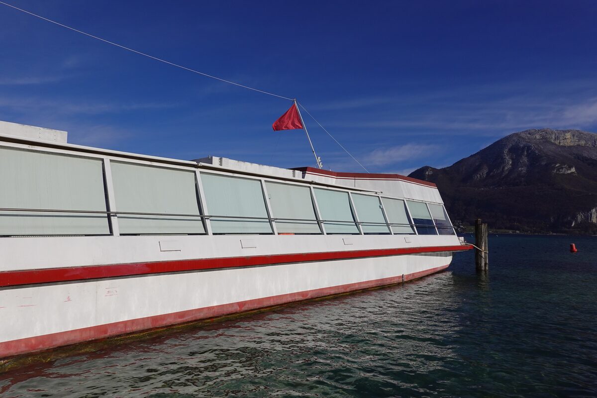 A white and red passenger boat with a red flag, tied to a wooden mooring post on Lake Annecy, with a rocky mountain across the water under a blue sky