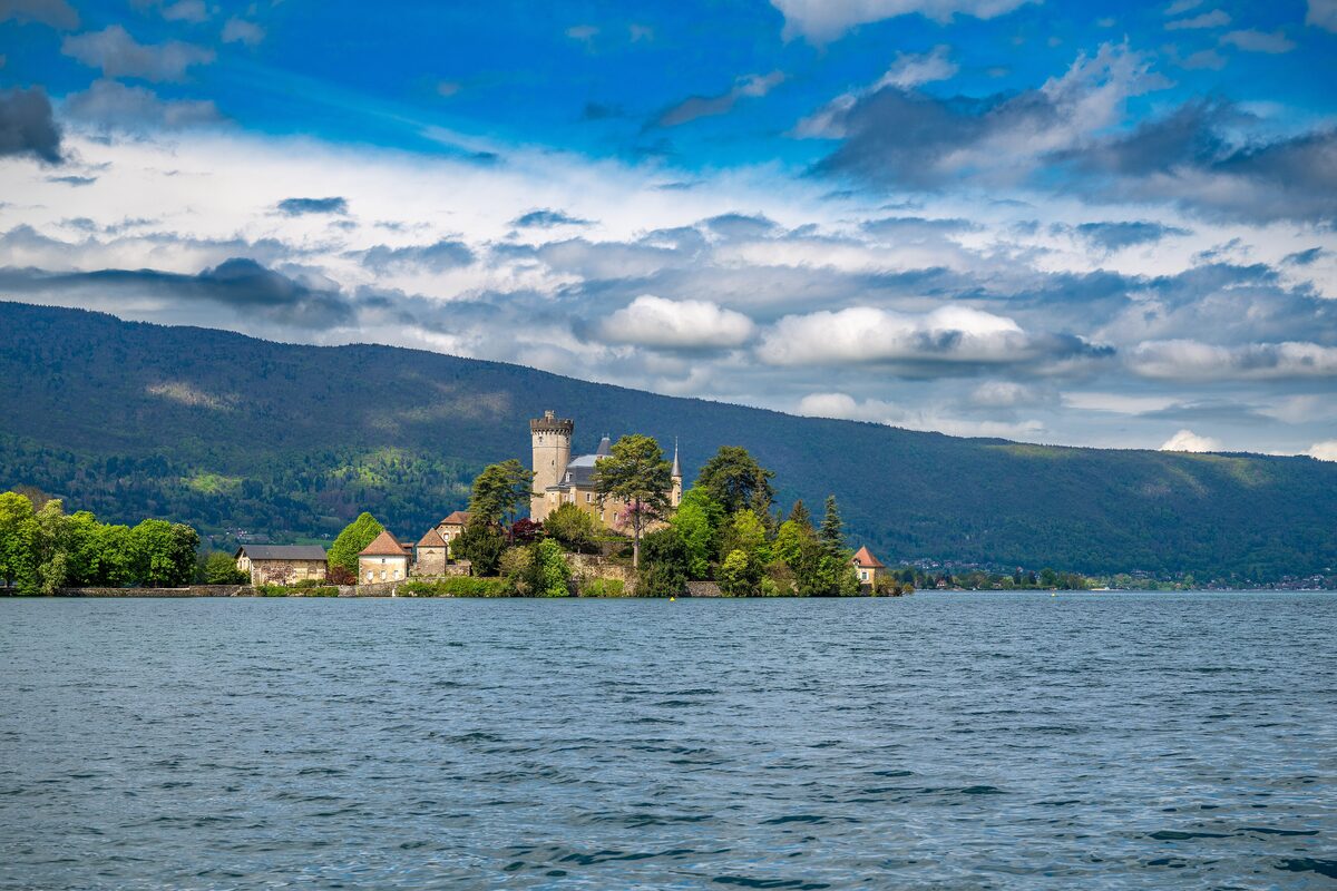 The Château de Duingt on its wooded point at the edge of Lake Annecy, a stone tower among trees and roofs under a blue sky with clouds