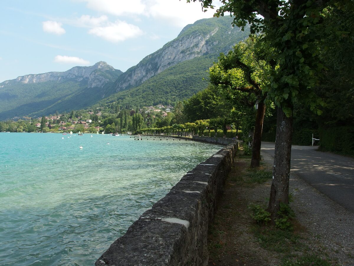 The Lake Annecy shore at Menthon, a stone quay wall and a row of trees beside turquoise water with the mountains behind