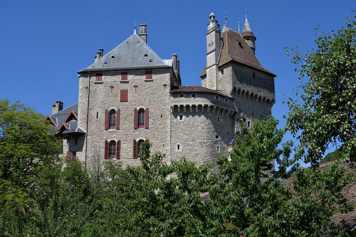 The Château de Menthon-Saint-Bernard, a stone castle with a tall tower and pointed turrets, half hidden by trees under a blue sky