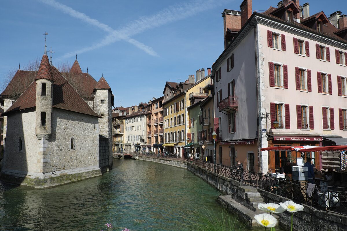 The stone Palais de l'Isle with its brown tiled turrets beside the Thiou canal, seen in daylight from the quay across the canal, with tall shuttered houses along the water