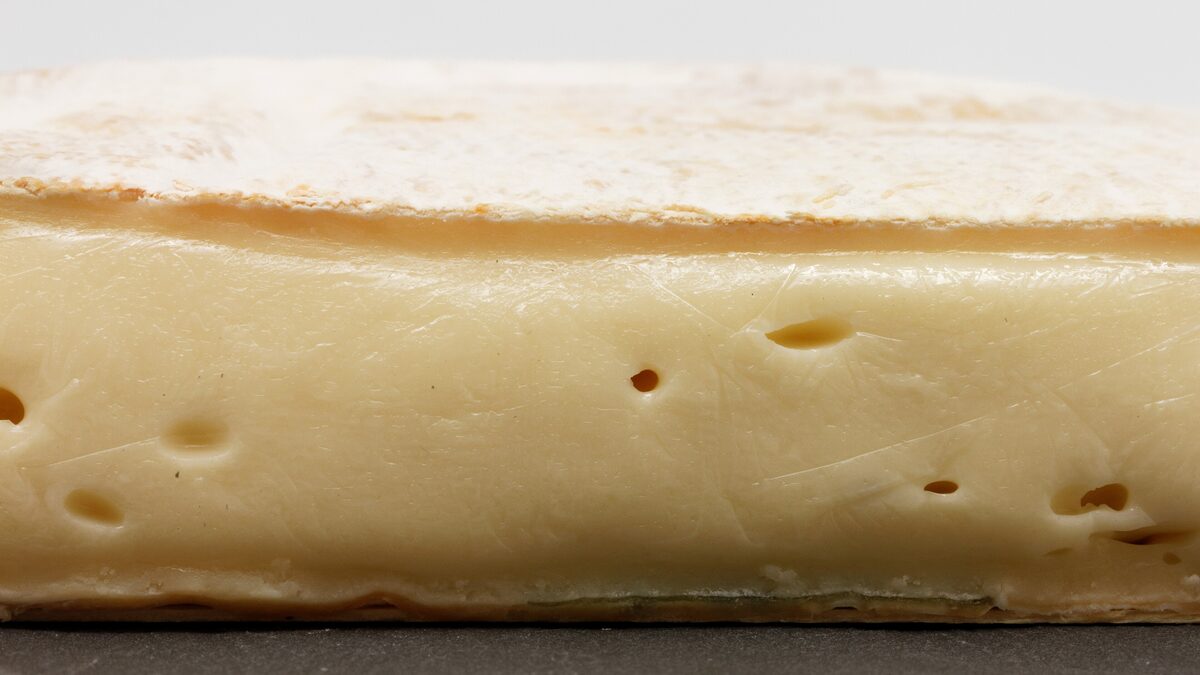 A cut Reblochon seen from the side, with its pale bloomy rind on top and a soft, creamy interior with a few small holes