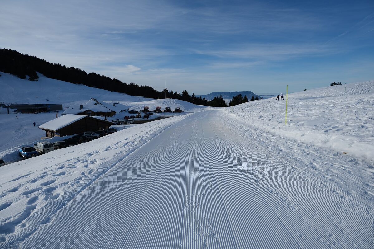 A groomed winter track on the Semnoz in snow, with a few low chalets and white slopes stretching across the ridge