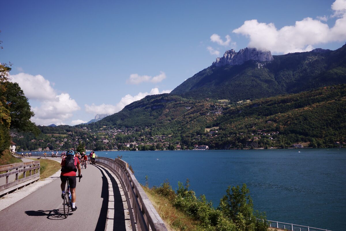 A cyclist on the lakeside Voie Verte, a wooden-railed path above the blue water of Lake Annecy with a limestone peak and the far shore beyond