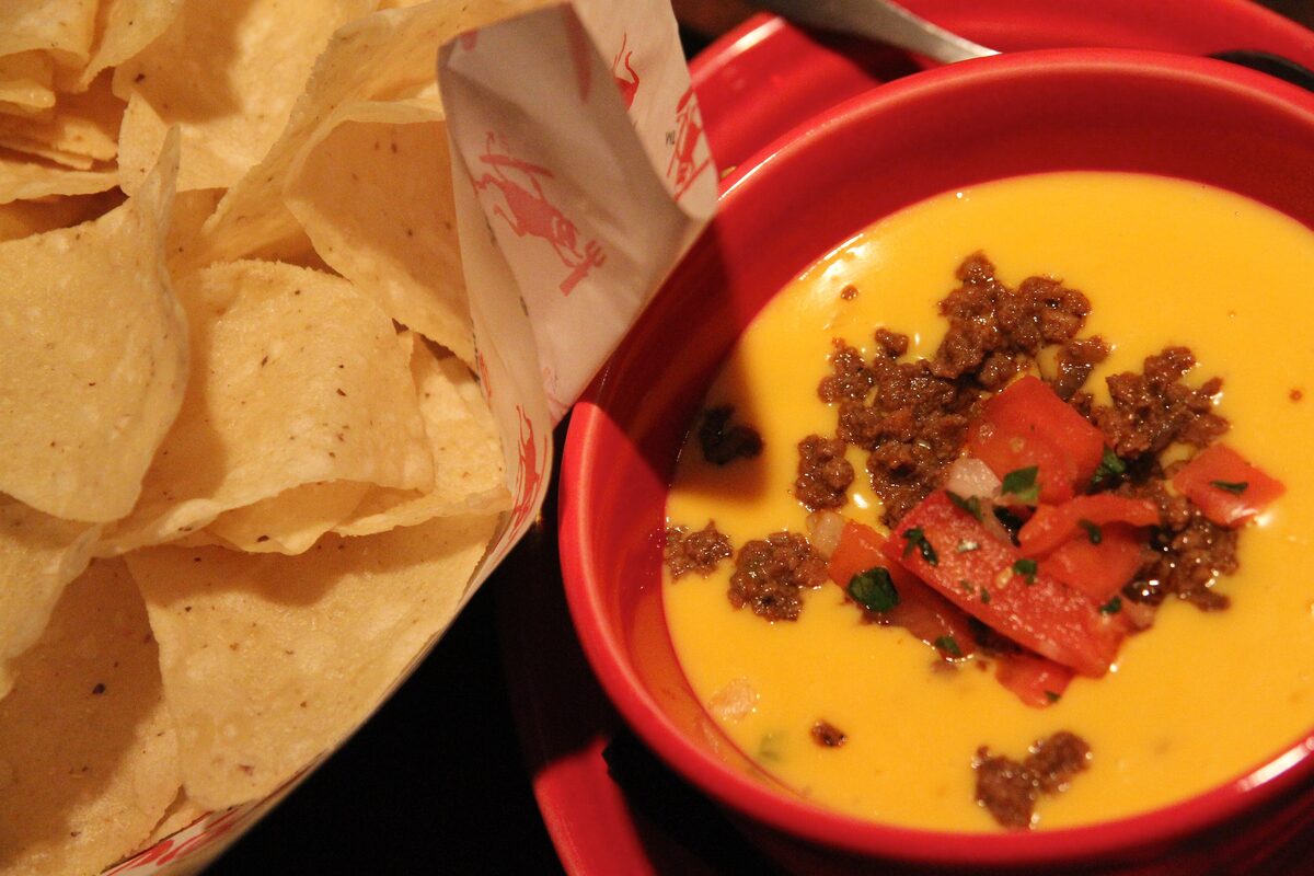 A red bowl of chile con queso topped with chorizo and diced tomato, next to a basket of tortilla chips
