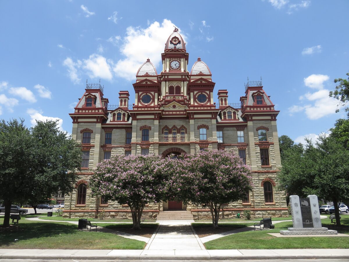 The red and cream Caldwell County Courthouse in Lockhart behind crape myrtle trees and a lawn