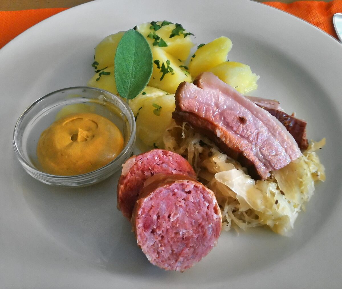 A plate of Berner Platte with sliced sausage and smoked pork on sauerkraut, boiled potatoes with a sage leaf and a dish of mustard