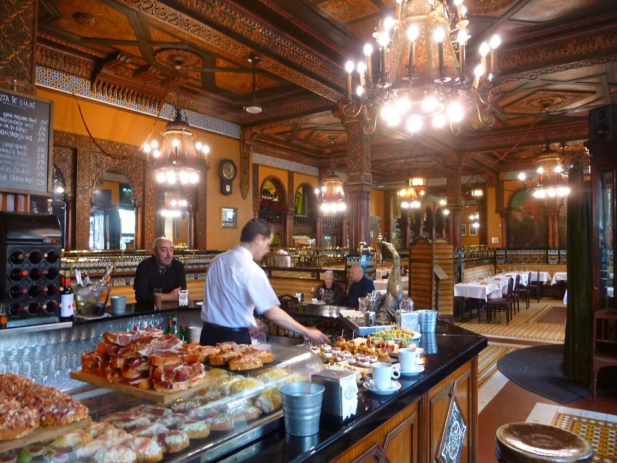 The ornate interior of Café Iruña, with a long bar, plates of food on the counter and chandeliers overhead