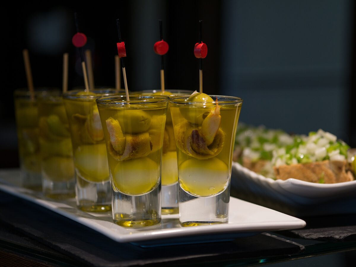 Small glasses each holding a skewered olive, anchovy and pickled pepper bite, served on a white tray