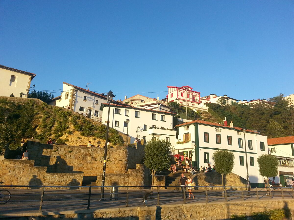 White and cream houses climbing a hillside above the old port of Algorta, with a stone wall and stairs below