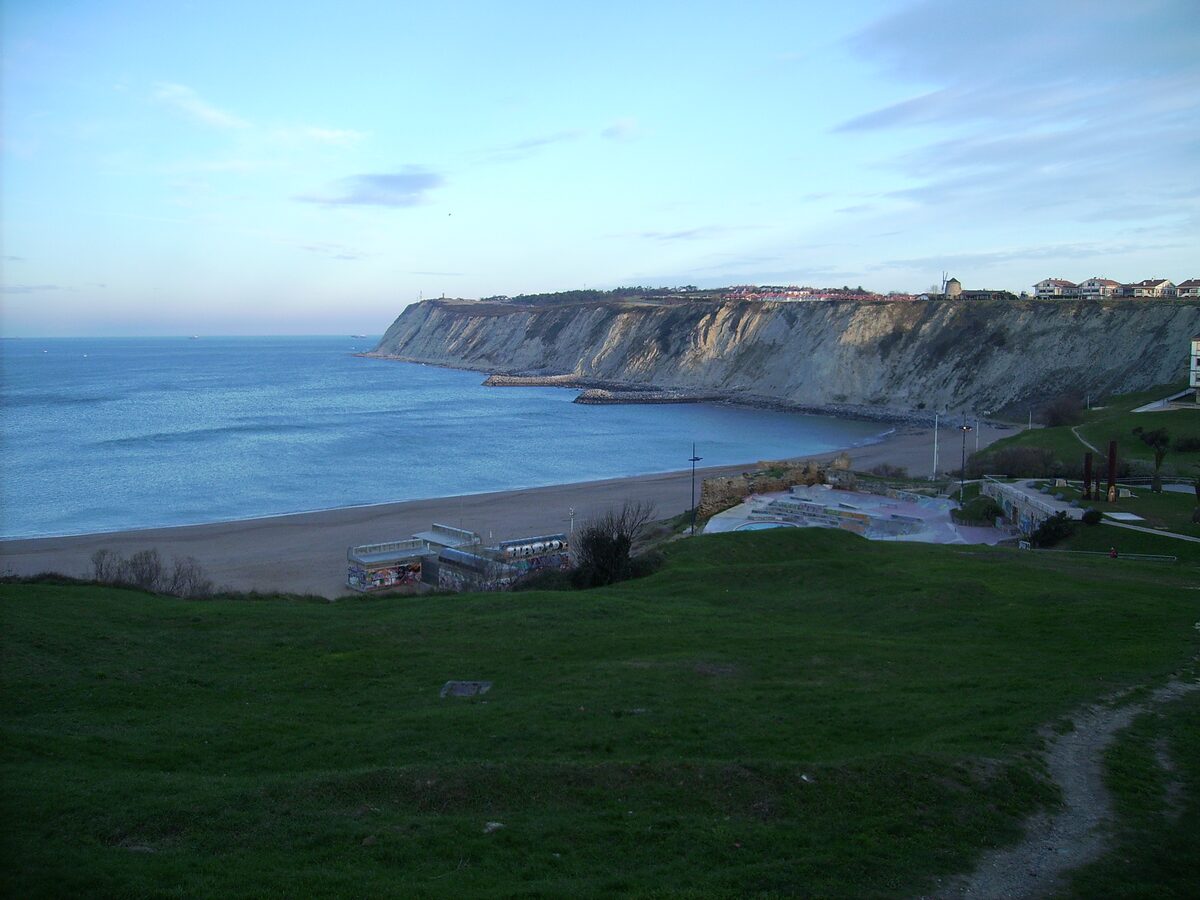 Pale cliffs of Punta Galea above a sandy beach and the Bay of Biscay, seen from a grassy slope at dusk
