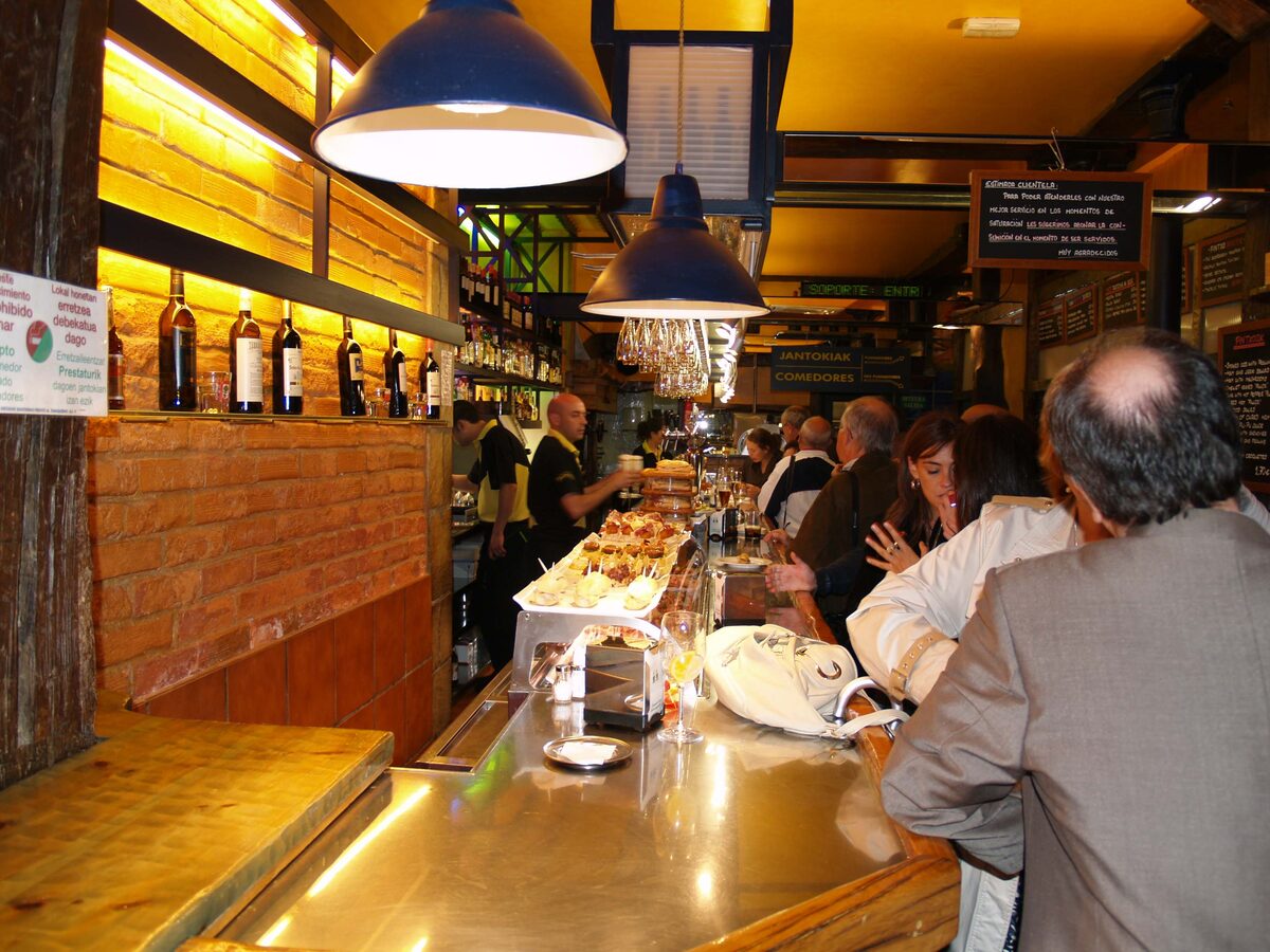 Customers at a busy Casco Viejo bar counter with pintxos under the glass and wine bottles on the wall