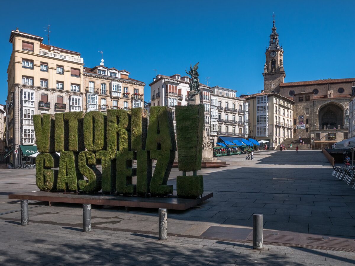 Green hedge letters spelling Vitoria-Gasteiz on the Plaza de la Virgen Blanca, with the church tower behind