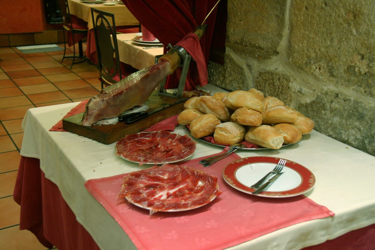 A ham on a stand beside plates of freshly sliced jamón and a plate of bread rolls in a stone-walled restaurant