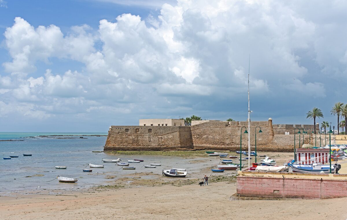 La Caleta beach under a bright sky, with small boats on the water and the walls of the Castillo de Santa Catalina behind